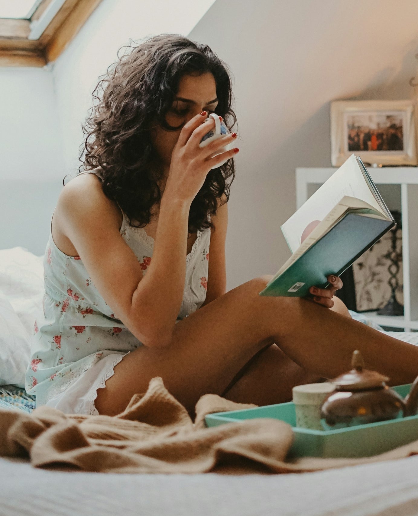 An image of a woman on her bed drinking from a cup and reading from a green book in the morning sunlight