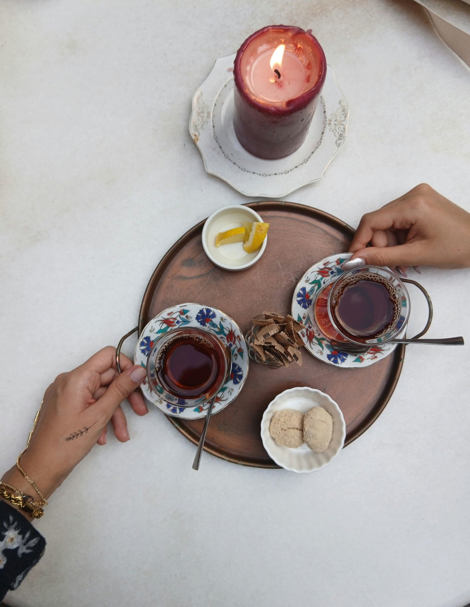 An image of two hands, each holding a cup of tea on a wooden tray, with a dark-coloured candle burning beside the wooden tray