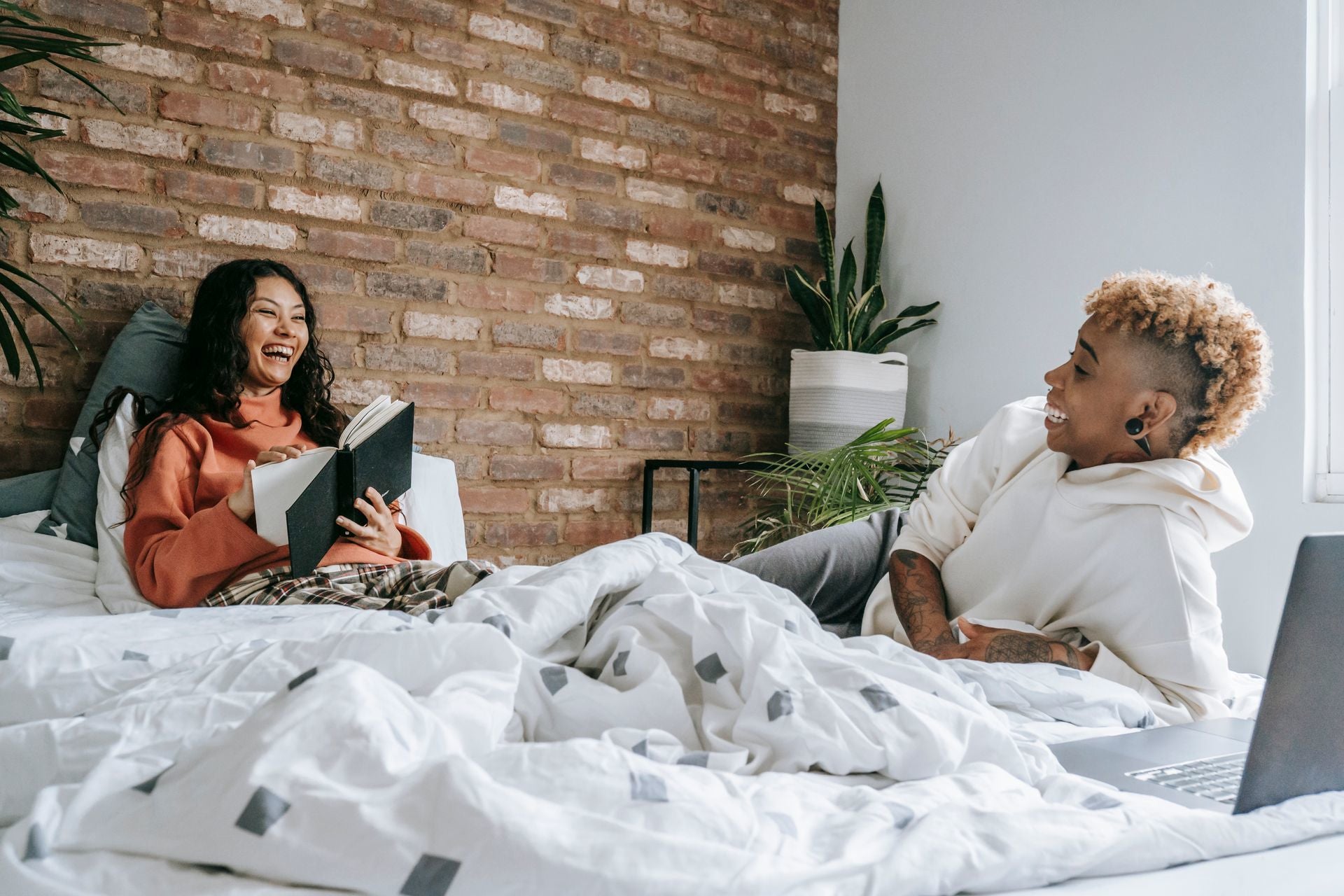 An inage of two woman laughing on a bed. The one women is holding a book.