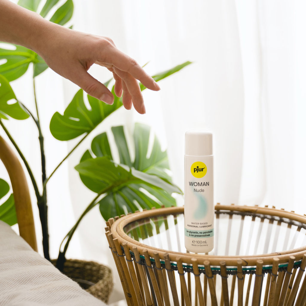 Hand reaching towards a bottle of pjur WOMAN Nude water-based lubricant on a glass and bamboo table with a plant in the background.