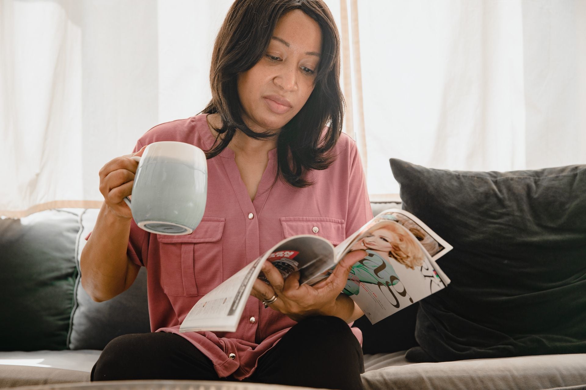 An image of a woman sitting on a couch in the morning light, holding a cup of something and reading a magazine